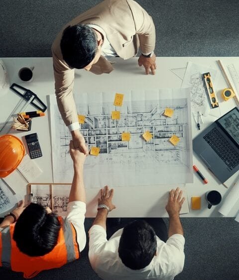 Top view of three professionals examining architectural plans on a large table with a laptop and tools, discussing Technology in Philippines Construction.