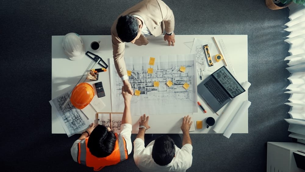 Top view of three professionals examining architectural plans on a large table with a laptop and tools, discussing Technology in Philippines Construction.