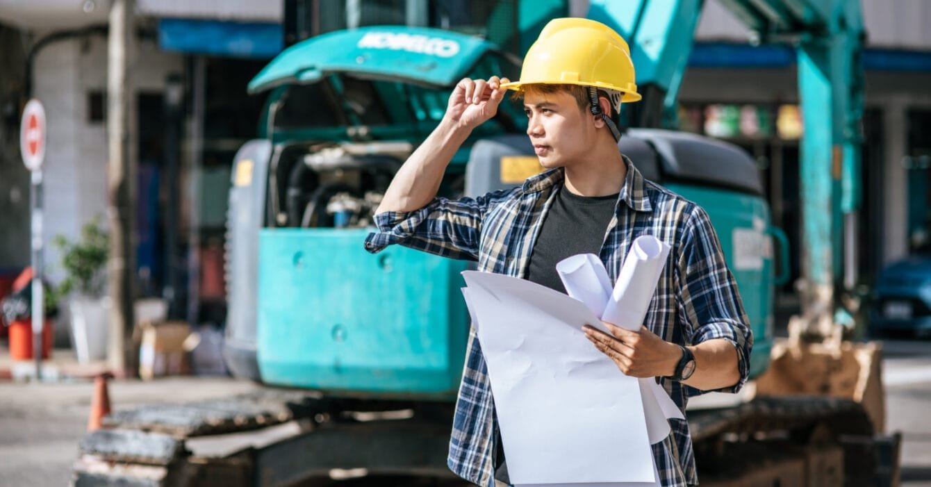 Infrastructure Development Plans Philippines: Construction worker with yellow helmet and blueprints at a site with an excavator in the background.