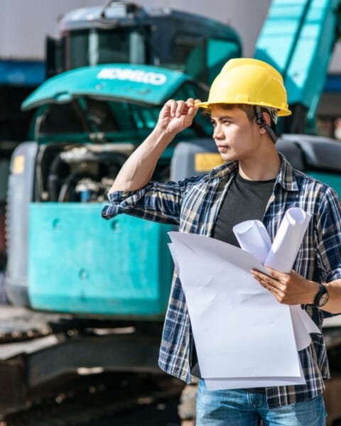 Infrastructure Development Plans Philippines: Construction worker with yellow helmet and blueprints at a site with an excavator in the background.