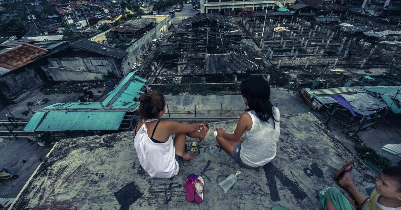 Shutterstock_1551064088 Two people sitting on a rooftop overlooking a densely packed neighborhood with damaged buildings, showing Philippines Housing Crisis.