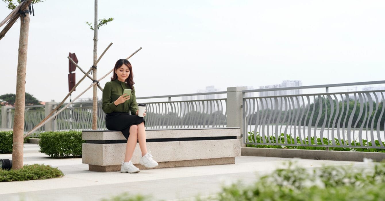 Philippines Sustainable Materials Usage: Woman sitting on a bench with a phone and coffee, overlooking a railing and greenery.