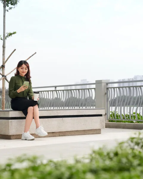 Philippines Sustainable Materials Usage: Woman sitting on a bench with a phone and coffee, overlooking a railing and greenery.