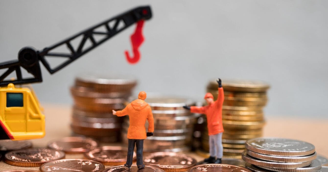 Philippines Infrastructure Investment Trends: Two miniature figures in orange jackets discussing near a pile of coins, with a toy crane overhead lifting a hook.