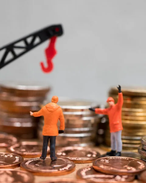 Philippines Infrastructure Investment Trends: Two miniature figures in orange jackets discussing near a pile of coins, with a toy crane overhead lifting a hook.