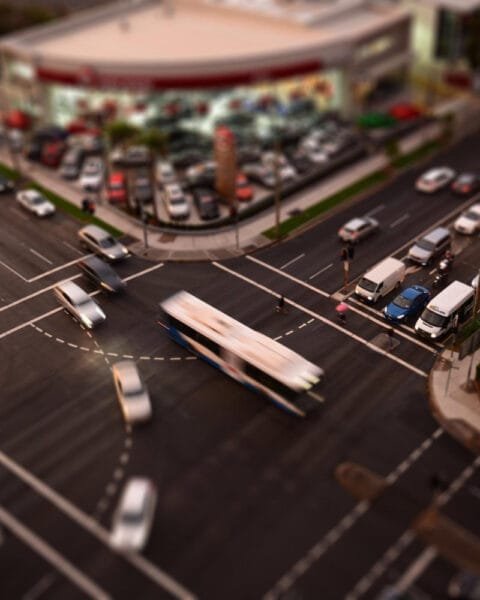 Aerial view of a busy urban intersection with blurred vehicles, including a bus and cars, near a dealership and tall buildings.