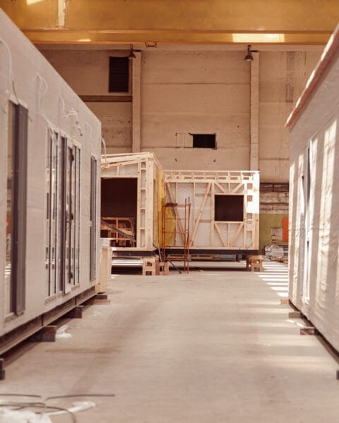 Interior of a factory showcasing partially built modular homes, with wooden frames and covered walls, arranged along a spacious aisle to show Philippines Modular Construction Adoption.