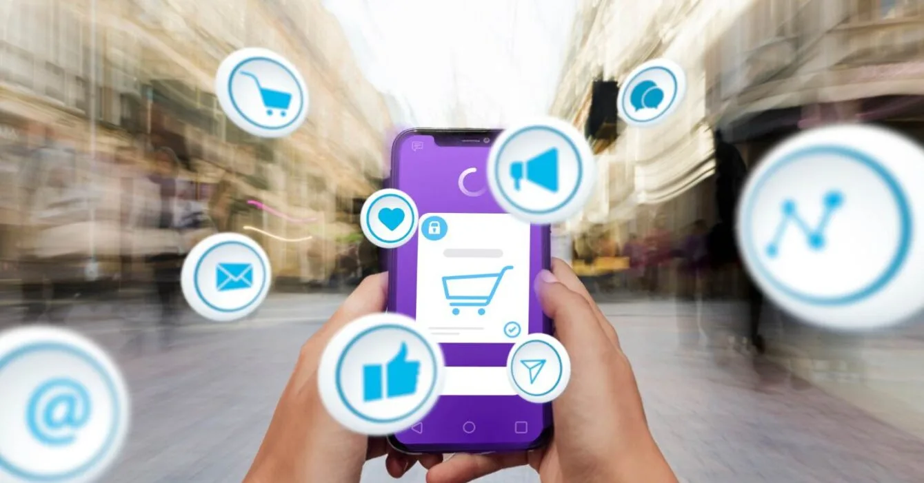 2150063168 Philippines Consumer Tech Trends: A person holds a smartphone displaying a shopping cart icon, surrounded by social media and communication symbols, in a blurred urban setting.