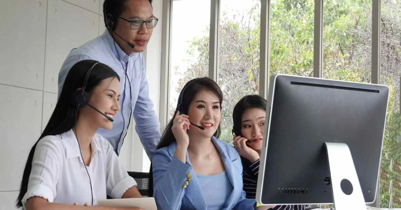 Philippines BPO Industry Outlook: A group of four people, wearing headsets, collaborate around a computer in a bright office space with large windows.