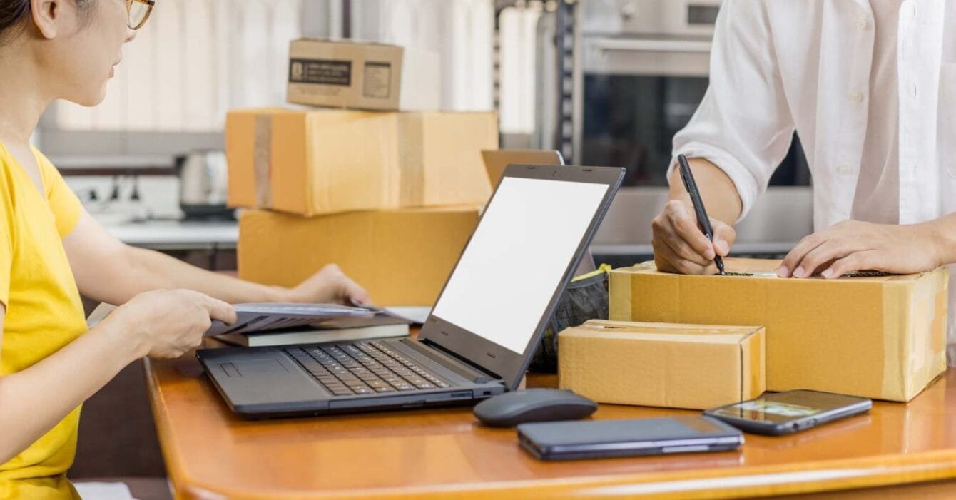 Philippines E-commerce Logistics Challenges: A person works at a wooden table with a laptop, writing on a box surrounded by cardboard packages and a smartphone.