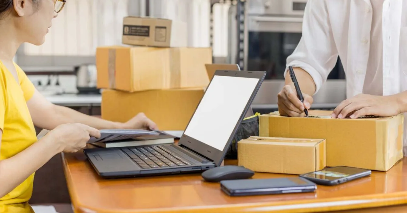 Philippines E-commerce Logistics Challenges: A person works at a wooden table with a laptop, writing on a box surrounded by cardboard packages and a smartphone.