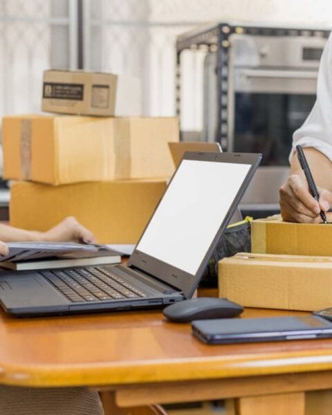 Philippines E-commerce Logistics Challenges: A person works at a wooden table with a laptop, writing on a box surrounded by cardboard packages and a smartphone.