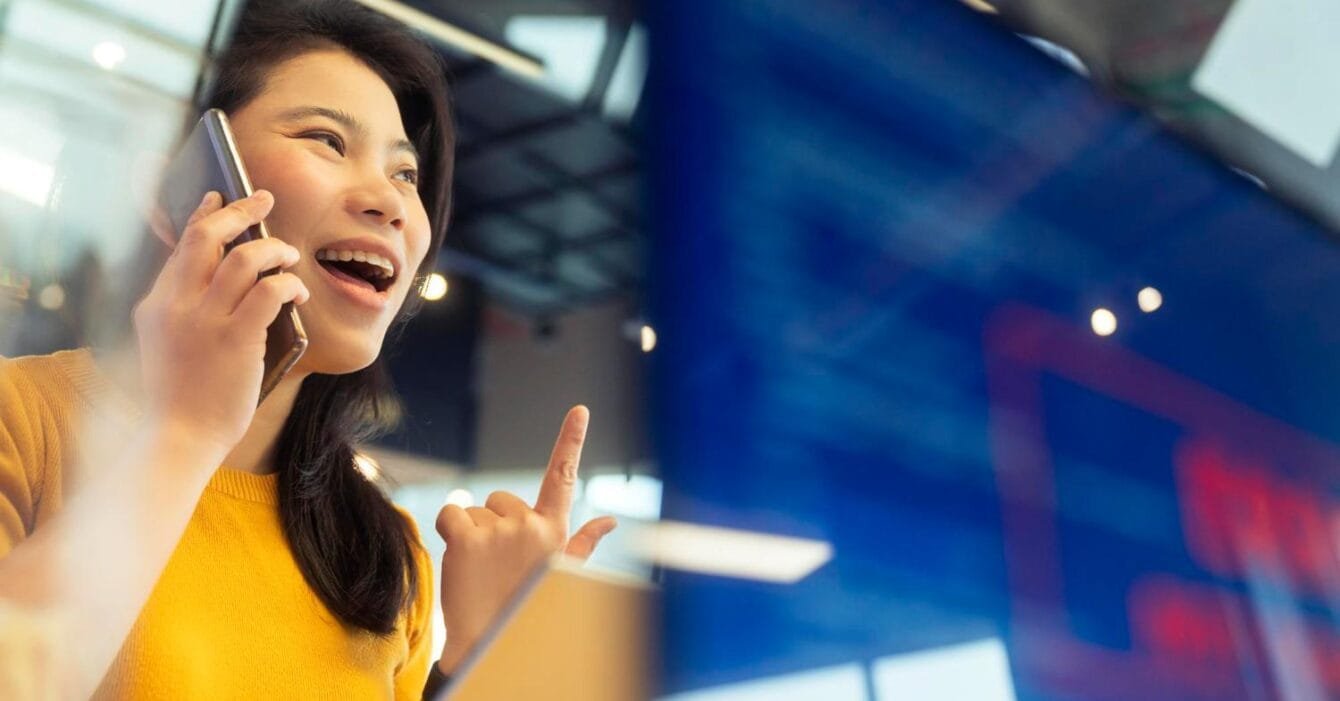 Philippines Telecom Consolidation: An Asian woman in a yellow sweater gestures while speaking on the phone in a modern, well-lit office space.