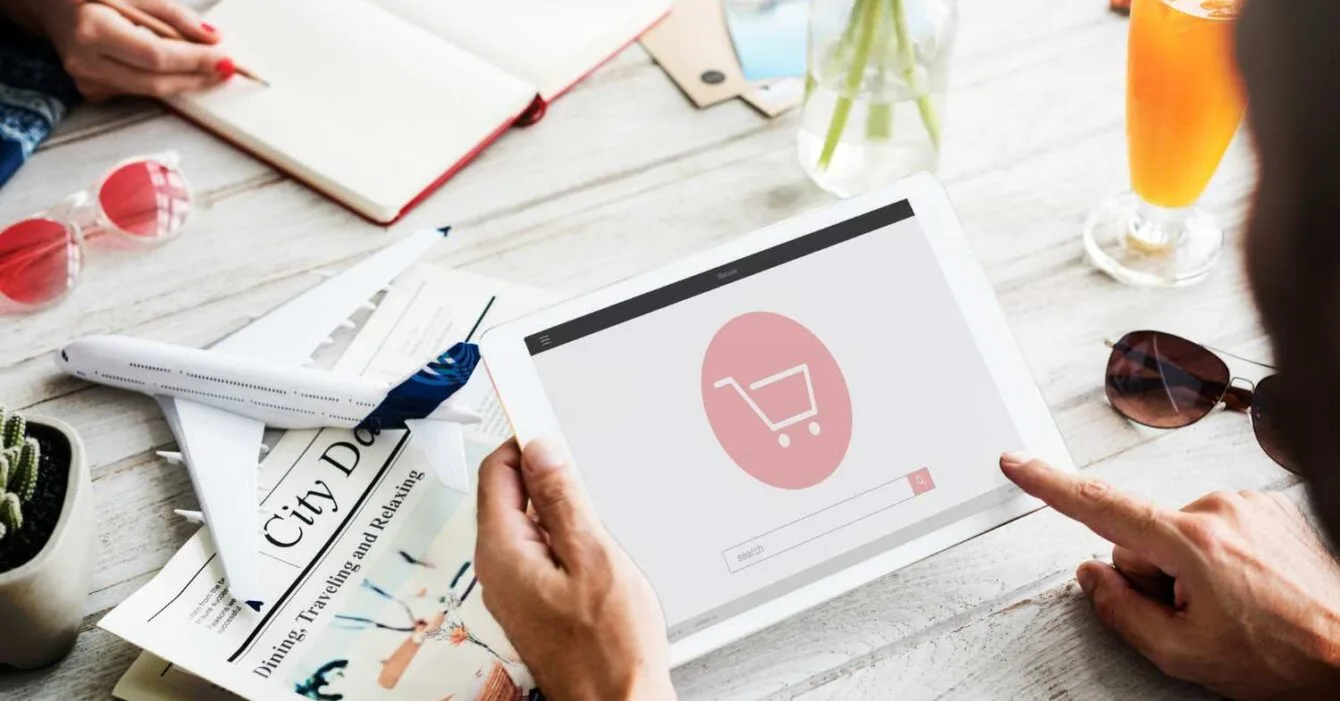 Philippines E-commerce Innovation: A person uses a tablet displaying a shopping cart icon while surrounded by travel-related items and a refreshing drink.