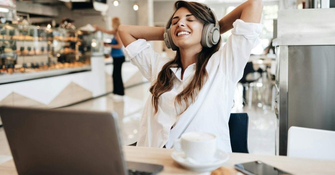 Freelance Economy Philippines: A woman in a white shirt relaxes in a café, wearing headphones, with a laptop, coffee, and a croissant on the table.