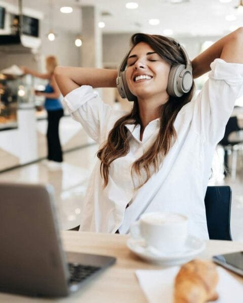 Freelance Economy Philippines: A woman in a white shirt relaxes in a café, wearing headphones, with a laptop, coffee, and a croissant on the table.