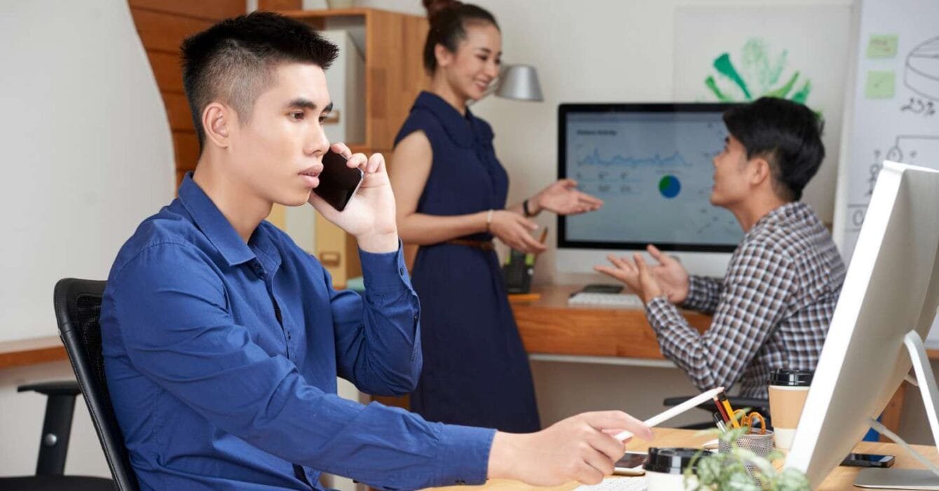 Philippines Outsourcing Transformation: A man in a blue shirt speaks on the phone at a desk; two colleagues engage in discussion near a computer displaying graphs.