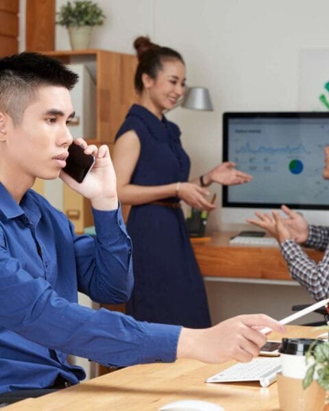 Philippines Outsourcing Transformation: A man in a blue shirt speaks on the phone at a desk; two colleagues engage in discussion near a computer displaying graphs.