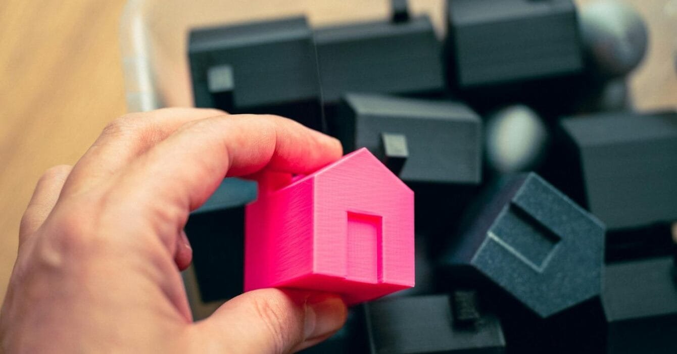 Philippines RE Price Concessions: A hand holds a bright pink 3D-printed house model above a container filled with black house-shaped models.