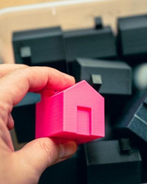 Philippines RE Price Concessions: A hand holds a bright pink 3D-printed house model above a container filled with black house-shaped models.