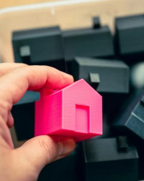 Philippines RE Price Concessions: A hand holds a bright pink 3D-printed house model above a container filled with black house-shaped models.