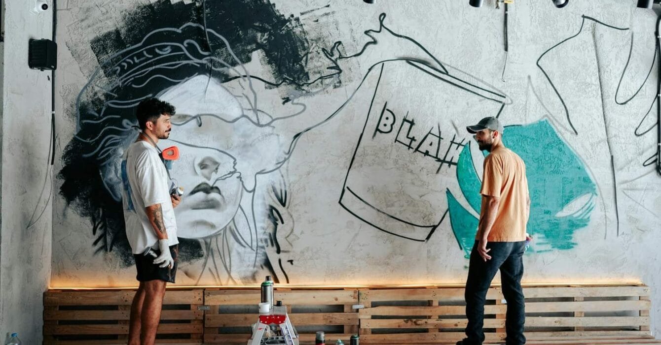 yunus-tug-PRI2vlMQiD0-unsplash Philippines creative economy 2026: Two artists work on a mural featuring a stylized face and abstract elements on a textured wall, surrounded by paint supplies.