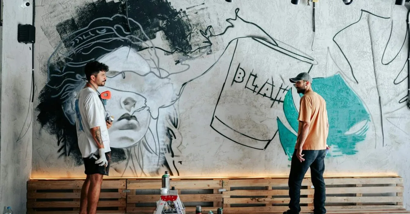yunus-tug-PRI2vlMQiD0-unsplash Philippines creative economy 2026: Two artists work on a mural featuring a stylized face and abstract elements on a textured wall, surrounded by paint supplies.